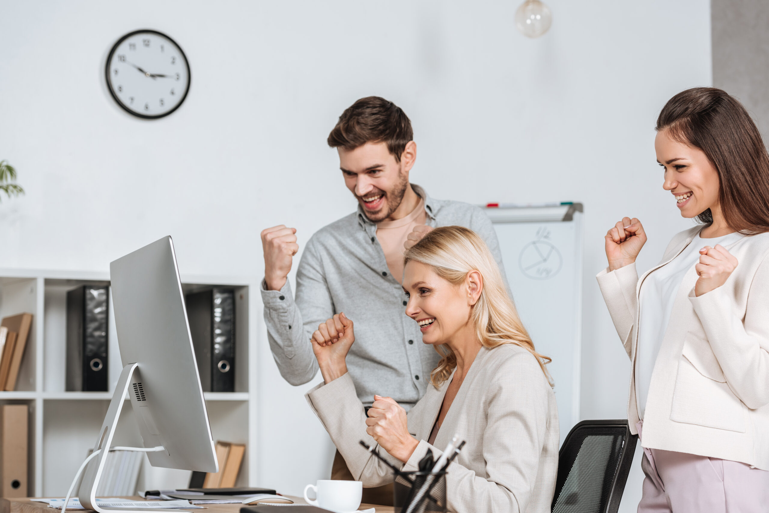 excited business mentor with colleagues shaking fists and looking at desktop computer in office
